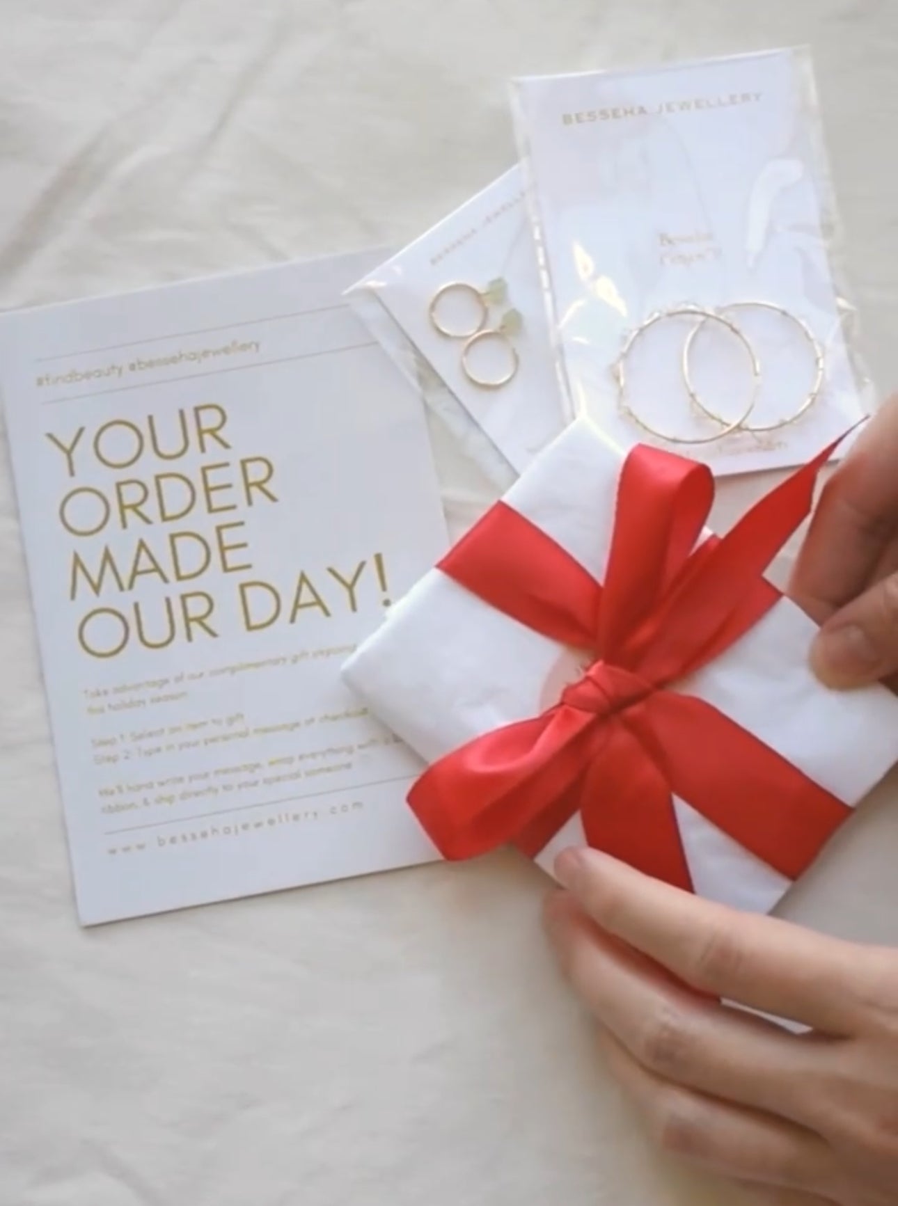 A person's hand is shown placing a red ribbon on a white gift wrap, with a printed message that reads 'YOUR ORDER MADE OUR DAY!' and a stack of gold-colored jewelry items.