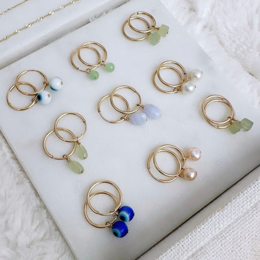 A collection of gold-filled hoop earrings with various colorful gemstone and pearl charms laid out on a white surface.