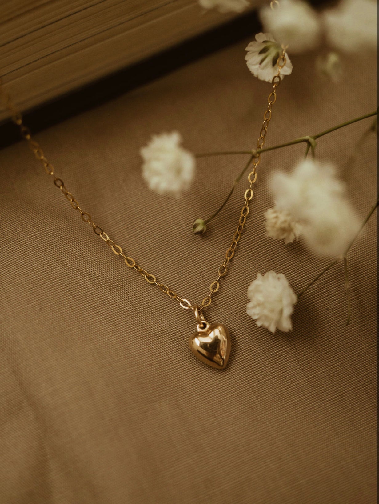 A gold-filled petite puffy heart necklace laid out on a fabric surface with small white flowers scattered around it.