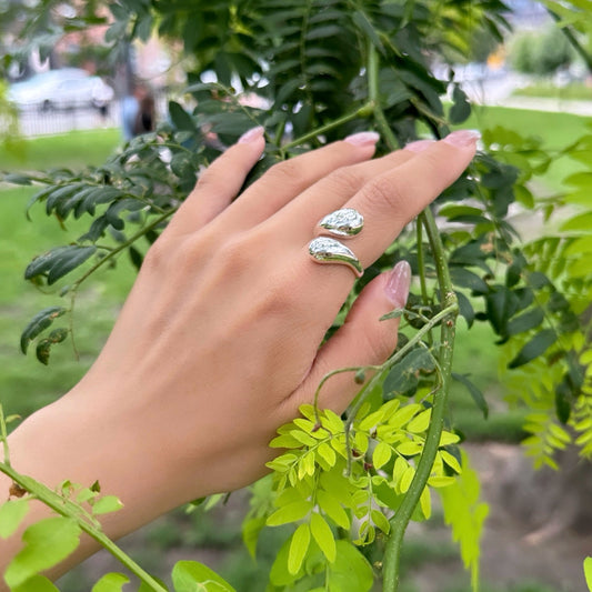 A person's hand wearing an adjustable sterling silver double drop ring, with the ring visible as it hovers over a leafy branch.