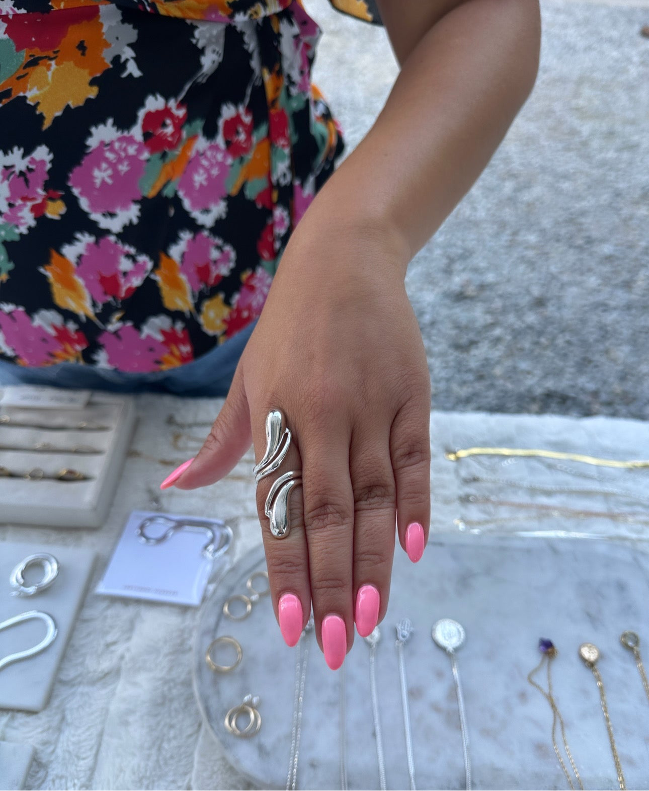 Hand wearing a silver ring with pink nails and floral-patterned sleeve, with jewelry on a marble surface.