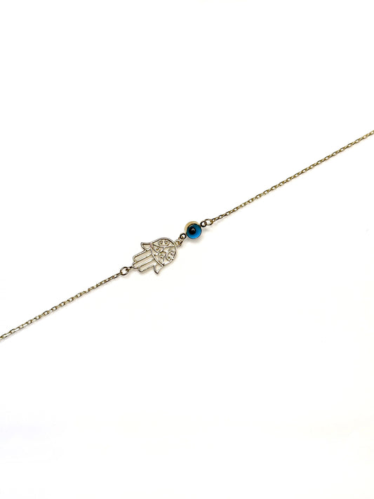 A gold bracelet featuring a Hamsa hand charm and a vibrant cobalt blue evil eye charm on a white background.