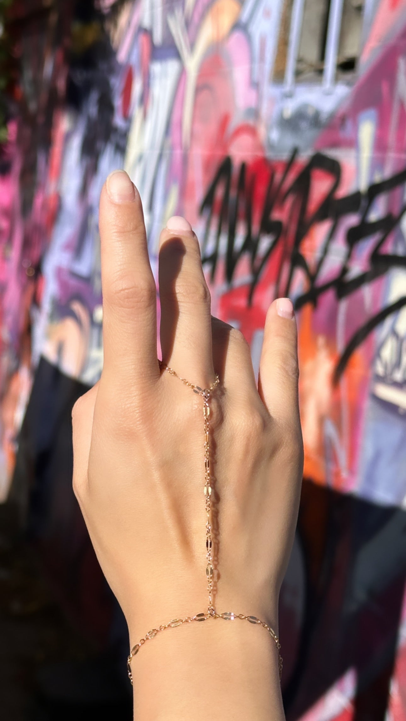 A hand wearing a gold-filled hand chain against a colorful graffiti background.