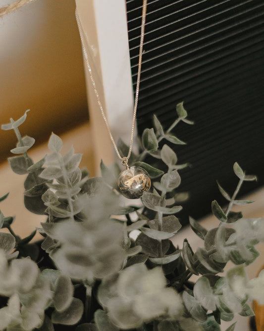 A sterling silver ball-shaped chime ball necklace with a chain, displayed against a floral background.