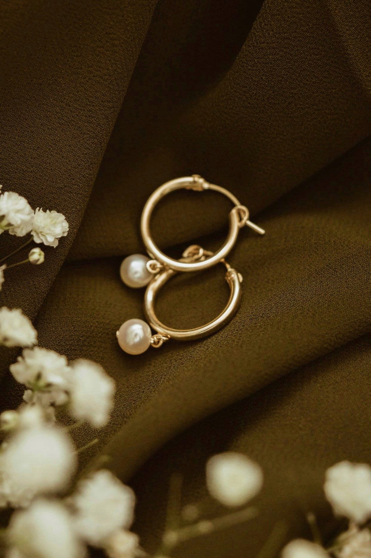 A pair of gold-filled hoop earrings with dangling freshwater pearls, displayed on a dark fabric background with small white flowers.