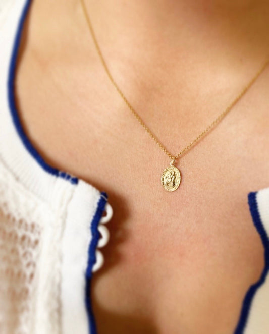 A close-up image of a woman wearing a petite St Christopher medallion necklace on a gold chain, displayed against a white garment with a blue trim.