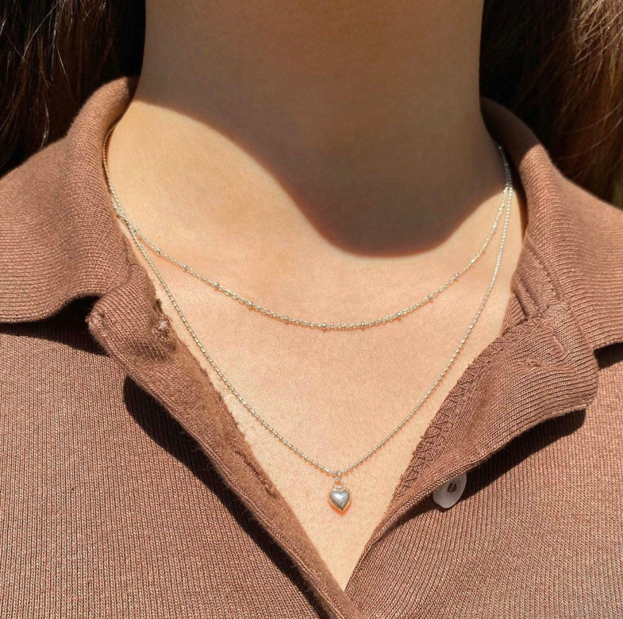 A person wearing a sterling silver petite puffy heart necklace on a slightly longer chain, positioned against a casual brown top.