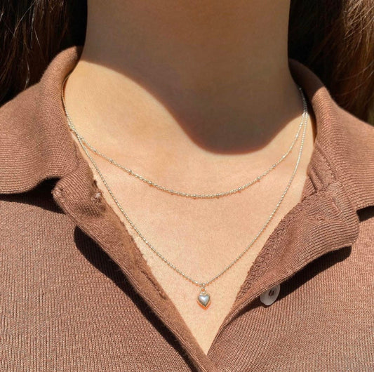 A person wearing a sterling silver petite puffy heart necklace on a slightly longer chain, positioned against a casual brown top.